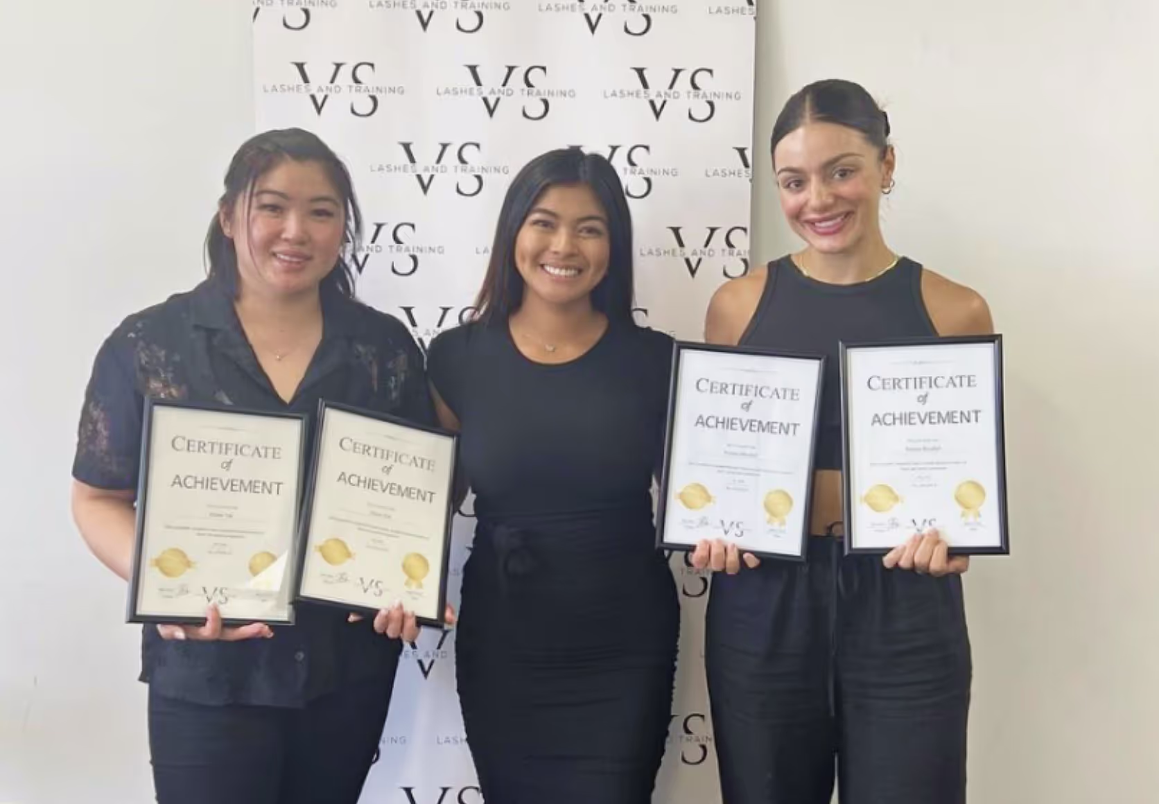 Trainer and students holding framed lash certification diplomas in beauty studio setting.