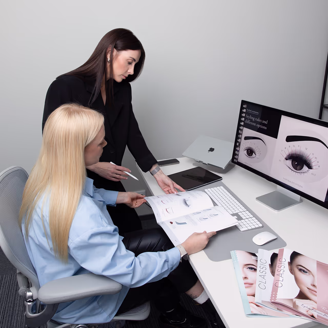 Trainer explaining lash training materials to a student at a desk with screen and printed manuals.