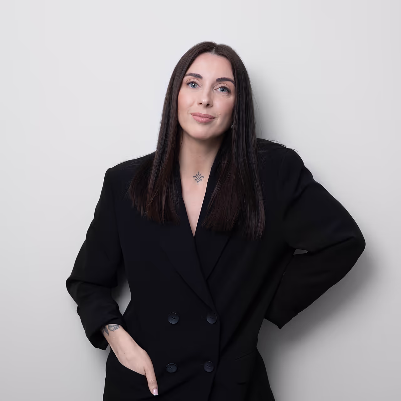 Woman with long dark hair wearing a black blazer, standing against a light gray background with a tattoo visible on her wrist and upper chest.