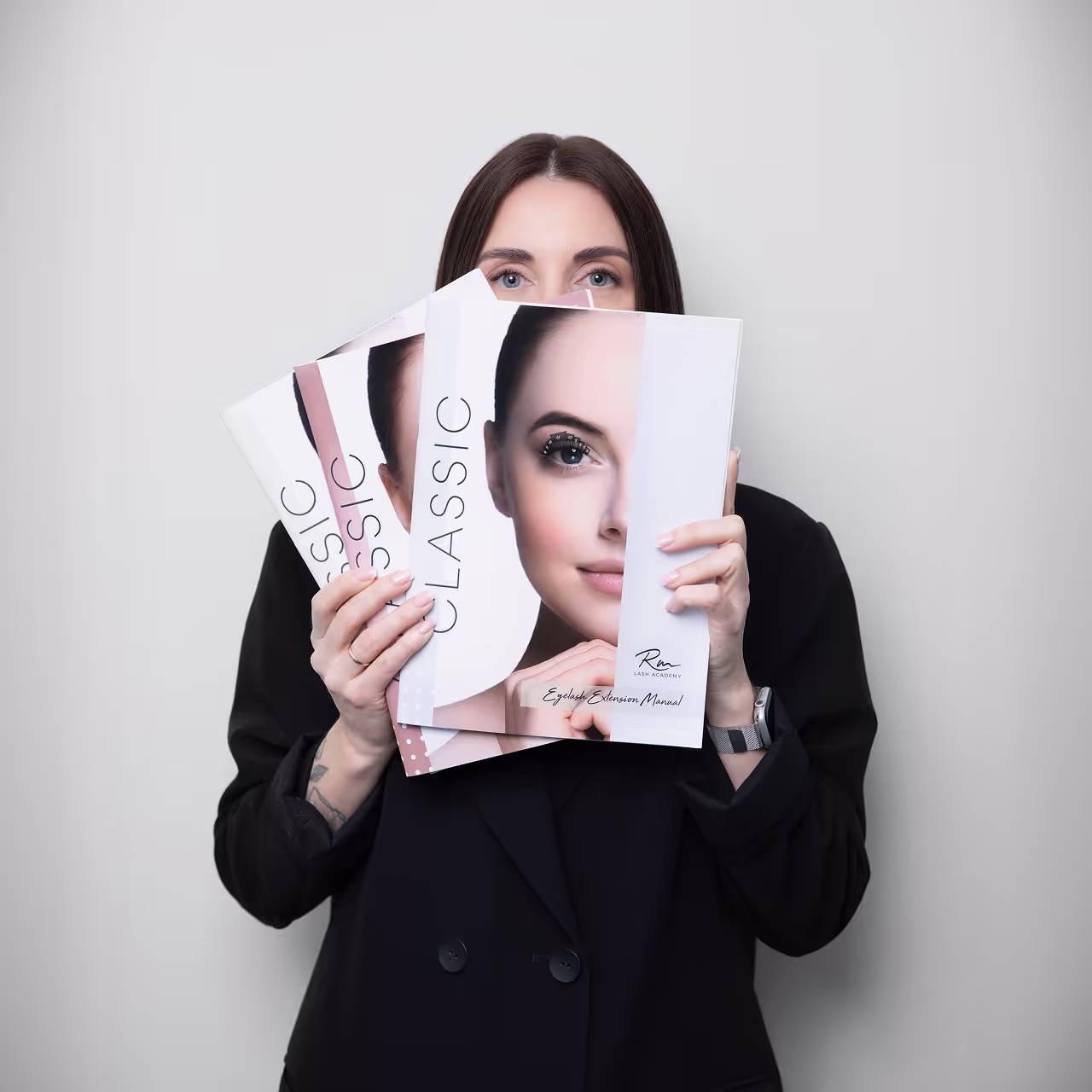 Woman holding RM Lash Academy eyelash extension manuals in front of her face, partially covering it.