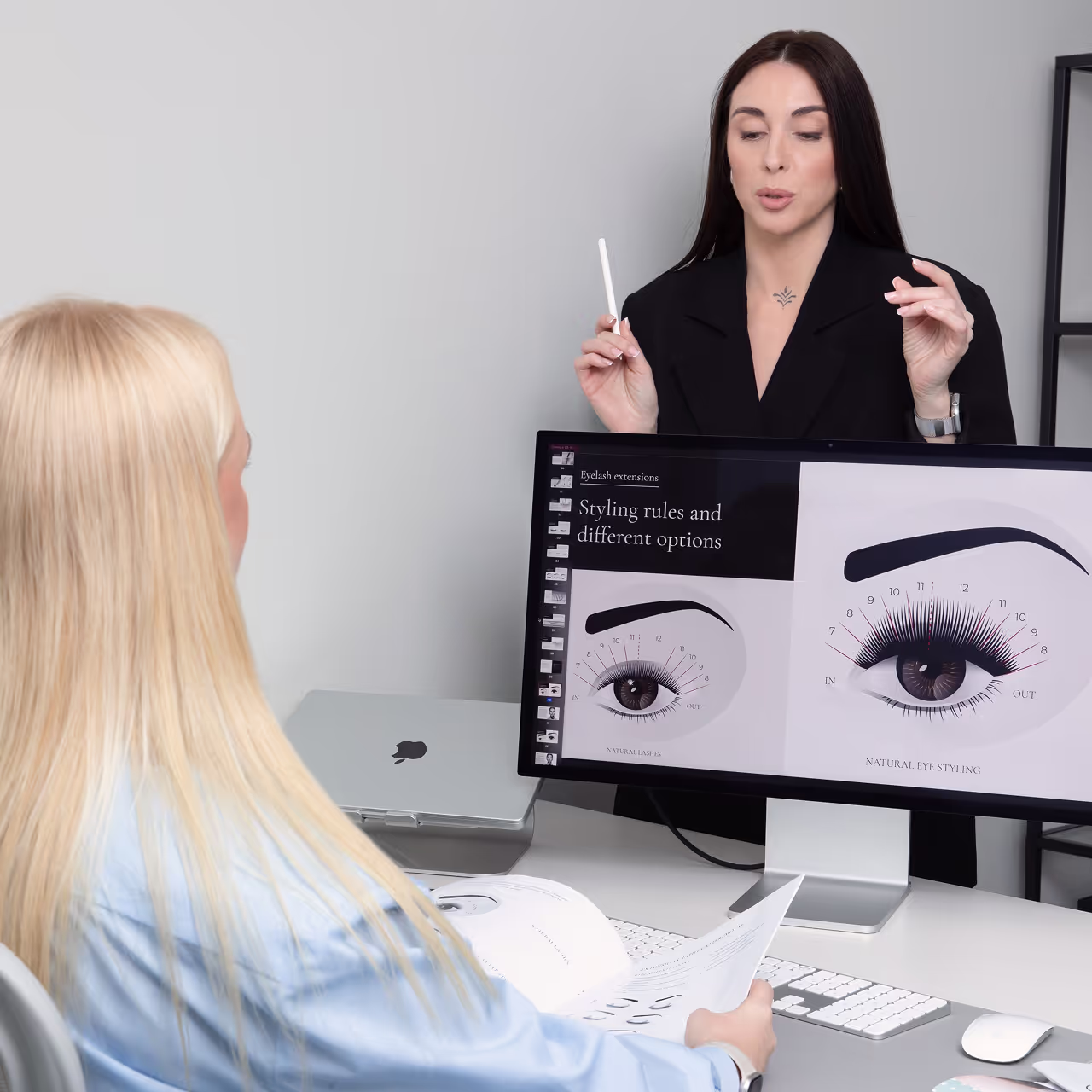 Lash training academy educator teaching a student about eyelash extension styling rules and eye shapes, using visual materials on screen and printed manuals.